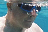 An older man under water in a pool exhales 2 bubbles through his nose.