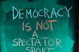 Green and black background with words in chalk writing “Democracy is not a spectator sport” — preventing democratic backsliding
