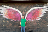 Mu’nis is a green shirt and blue jeans, standing in front of wall art depicts red and orange angel wings.
