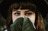 A woman with dark bangs and grey-green eyes. She is wearing a black hat and is holding a large leaf in front of the lower half of her face.