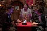 The white mother and her two teen boys look at a row of lit-up gemstones on the table in front of him. They’re all raising their hands excitedly, reacting to the gemstones lighting up