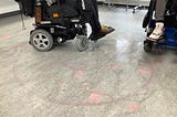 An office floor with chalk tire marks in two perfect circles. The artist is visible in his powered wheelchair.