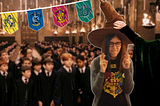 Maayan, the writer of this article, a young woman with glasses and a Hogwarts t-shirt, crossing her fingers at a Hogwarts sorting ceremony, wearing a sorting hat