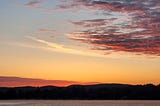 photograph of sunrise, with most of the image being the sky and clouds, though a bit of the horizon and the lake beneath is visible, with wispy fog in the distance just over the water