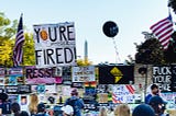 Anti-Trump activists with signs in a photo from 2020 in Washington, DC, saying “You’re Fired,” “Resist,” and “Fuck Your Fence.”