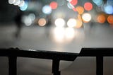 Hand rail with blurry street behind it at night with lots of lights. Part of the image is contained within a circle and rotated slightly to break the line of the hand rail.