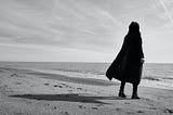 Greyscale image. Person in a black cloak stands alone on a beach near the edge of the water looking out, their shadow extending behind them.