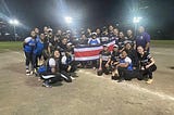 Photo of the Whittier Softball team alongside the team in Costa Rica, holding the Costa Rican flag on the softball diamond.