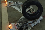 A tire sits on the tip of a v-shaped pile of newspaper. 2 flames on either side are just beginning to ignite the newspaper.