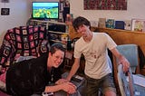 A photo of the author leaning over a padded footrest covered with a waterproof sheeting and wearing a TENS machine. They are smiling and their partner is holding their arm and smiling too. There are sunflowers on the TV behind them and other birth detritus is scattered around the room.