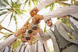 Low angle photo of half-a-dozen white men clinking their beer bottles together. It’s daytime with a sunny sky and palm trees overhead, and the men are wearing sunglasses and suits with designer stubble.