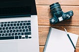 Flatlay image of a desk with a laptop, notebook, and camera arranged neatly on it.