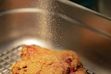 a piece of fried chicken getting peppered in a basket.