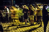Time-lapse photo of Pokémon Go players in Lillo (Antwerpen) from November 2016. The photographer must have played with effects, because there’s a beautiful YELLOW cast over the cobblestone street and across the players :). To the right-center, there’s a loose group of three teen boys watching another player who’s just walked past them, right to left. The person is just a blur, meaning they’re moving FAST, and the boys look a bit anxious, like there’s something they’ve yet to figure out :).