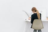 A woman sitting at a desk, facing the wall, writing in a journal.