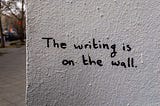 Muro branco com texto em inglês escrito “The writing is on the wall”