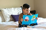 A young boy sits cross-legged on a neatly made bed, reading a bright blue children’s book. He wears brown pajamas with dark blue pants patterned with small animals. Behind him are white pillows with leafy designs and a purple accent pillow.