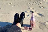A pink bottle of champagne, heart-shaped sunglasses and a girl’s feet in combat boots on the beach.