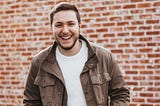 A young man smiling, in front of a brick wall.