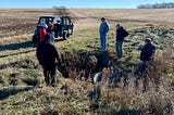 Six people stand near the edge of a field and circled around an outlet pipe.