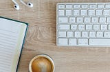 Fotografia com vista superior do tampo de uma mesa de madeira, onde aparecem: o canto superior de um caderno aberto com folhas pautadas, na parte superior da fotografia há fones de ouvido branco e um teclado de computador com teclas claras entre branco e cinza, proximo ao caderno há uma xicara de café.