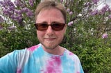 The author is wearing trans colors and standing in front of a flowery bush. He’s wearing sunglasses and has short brown hair with a mustache and short beard. He’s smiling slightly.