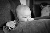 black and white. baby boy leaning over the edge of an arm chair, looking off into the distance.