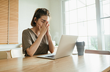 Image of woman sitting at a table in front of a computer with her hands on her face and eyes closed, apparently frustrated or worn out.