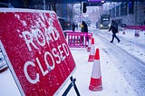 A sign viewed from its side says “Road Closed” in snowy weather next to traffic cones, a frozen road, pedestrians walking by, and a bus arriving.