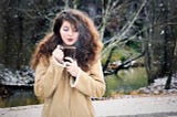 Girl, long brunette hair, winter coat, background woods and a river, blows steam off a cup of coffee