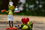 Ceramic frog chef holding a covered tray beside a frog lying at her feet holding a big red heart.