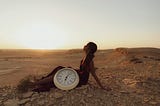 A woman is sat in a desert at sunrise with a large clock next to her