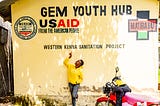 A smiling woman in a yellow sweatshirt stands next to a red motorbike in front of a wall and points to painted words on the wall that say: “Gem Youth Hub USAID. From the American People. Western Kenya Sanitation Project.”