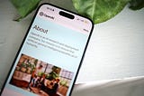 A phone with the “About” section page for OpenAI. The phone is on a white table, with leaves from a plant also in frame.