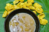 A black bowl with brownish yellow liquid featuring granules is placed over a banana leaf, yellow coloured jackfruit bulbs surround the bowl.