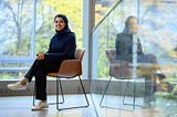 Sarah Alnegheimish sits in front of a wall of windows overlooking out-of-focus trees.