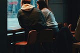 Two people sitting side by side in a café, looking out the window, suggesting quiet tension and emotional distance.