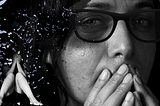 women with slight bruise near eye with tear over it hidden under dark sunglasses. In the corner under shattered glass a woman sits hiding her face.