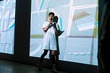 lauren holds a mic standing in front of a projection of casey’s artwork, featuring shapes of different colors rendered as lines overlapping