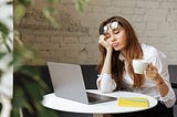 Tired woman sits with eyes closed at a computer, holding coffee