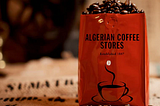 Image of Red Coffee Bag full of coffee beans — Bag printed “Algerian Coffee Stores Established 1887”, sitting on a coffee sack labelled SUMATRA