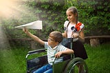 Girl with flowered blouse pushing a young boy in wheelchair, he is about to throw a paper airplane.