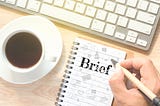 Picture of s hand writing “Brief” on a notepad next to a keyboard and mug of coffee