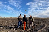 Three people stand around a bright orange drain inlet in a harvested field where construction finished recently, revealing black soil.