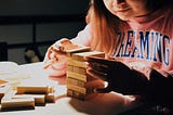 A child in a pink jumper that reads “dreaming” stacks wooden blocks on top of each other on a white desk.