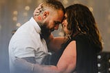 A woman in a black gown embraces a tattooed man in a white shirt
