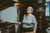 An older woman in the kitchen laughs as she holds a metal spatula next to the fryer