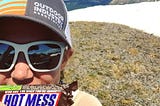 Author standing on mountain top, holding a Snickers bar that says “hot mess” on label