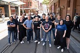 A group of people standing on tram tracks in front of red brick buildings, smiling. CyRise Accelerator startup founders with CyRise team. Back: Ron Moritz, Simon O’Keefe, Sam Stewart, Ivan Vanderbyl, Tony Mao, Shan Kulkarni, Tim Thacker and Fiona Long. Front: Tinesh Chhaya, Kirstin McIntosh, Lorenzo Modesto, Lainie Vinikoor, Scott Handsaker and Jenny Binetter