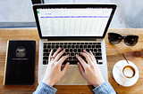 A person’s hands are typing on a laptop on a wooden desk. A spreadsheet is on the screen. A cup of coffee, sunglasses and a notebook are nearby on the desk.